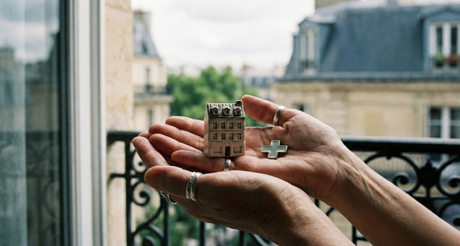Hands holding a miniature Parisian home and green medical cross representing health insurance in France for foreigners.